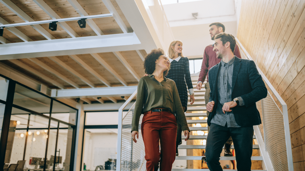 Four diverse professionals walking down an office staircase, smiling and engaging in conversation, representing a collaborative and positive workplace environment.