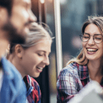 A group of three young adults, two women and one man, are laughing together outdoors. The woman in the foreground on the right, wearing a plaid shirt and glasses, is smiling broadly with her mouth open and appears to be speaking or gesturing.