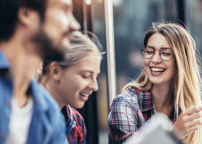A group of three young adults, two women and one man, are laughing together outdoors. The woman in the foreground on the right, wearing a plaid shirt and glasses, is smiling broadly with her mouth open and appears to be speaking or gesturing.