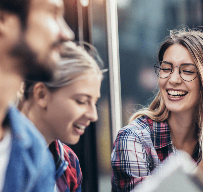 A group of three young adults, two women and one man, are laughing together outdoors. The woman in the foreground on the right, wearing a plaid shirt and glasses, is smiling broadly with her mouth open and appears to be speaking or gesturing.