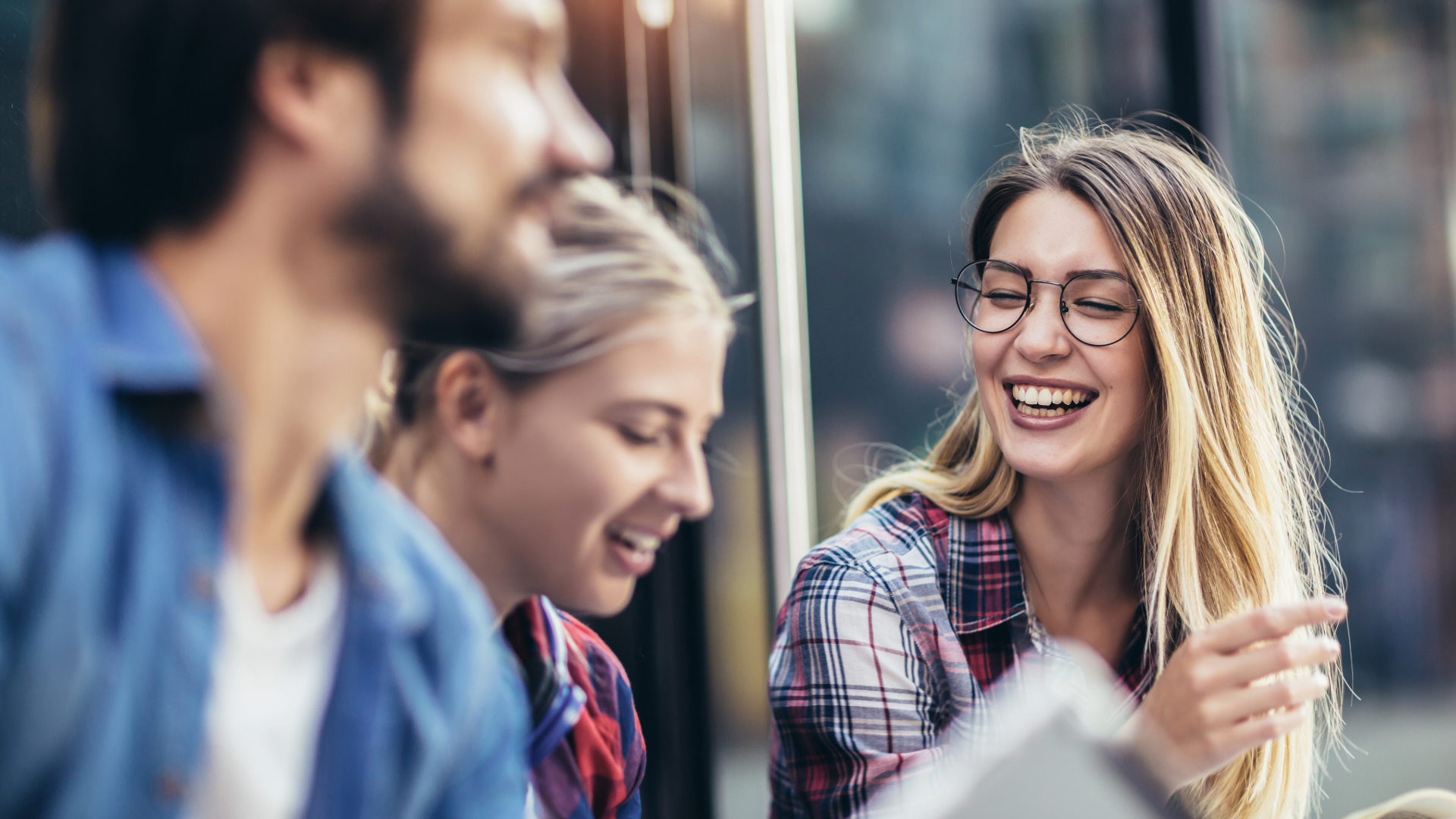 A group of three young adults, two women and one man, are laughing together outdoors. The woman in the foreground on the right, wearing a plaid shirt and glasses, is smiling broadly with her mouth open and appears to be speaking or gesturing.