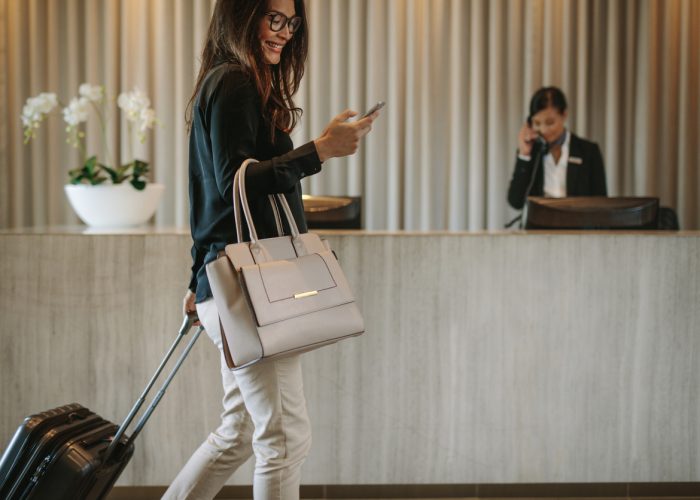 Woman using mobile phone and pulling her suitcase in a hotel lobby. Female business traveler walking in hotel hallway.