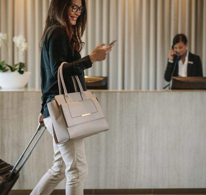 Woman using mobile phone and pulling her suitcase in a hotel lobby. Female business traveler walking in hotel hallway.