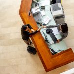 Overhead view of two women interacting at a modern reception desk with a light-colored top and wood paneling.
