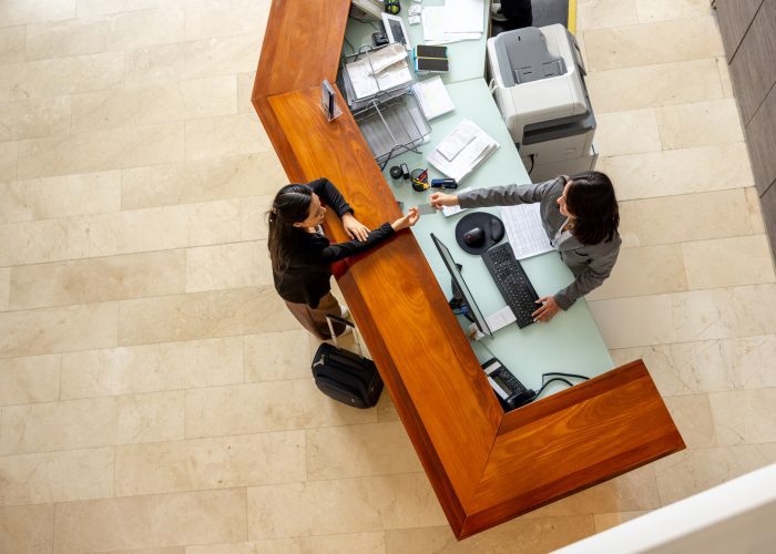 Overhead view of two women interacting at a modern reception desk with a light-colored top and wood paneling.
