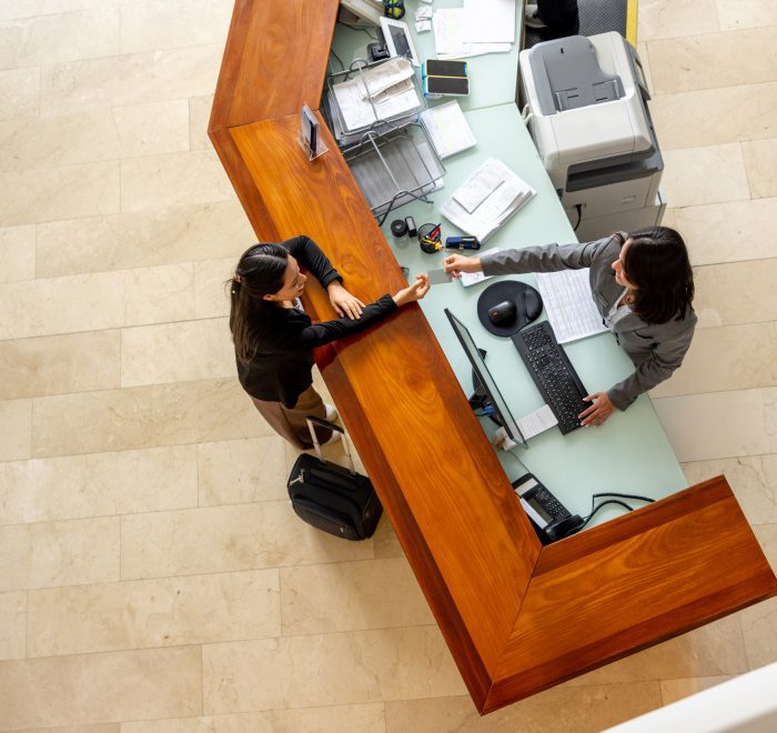 Overhead view of two women interacting at a modern reception desk with a light-colored top and wood paneling.