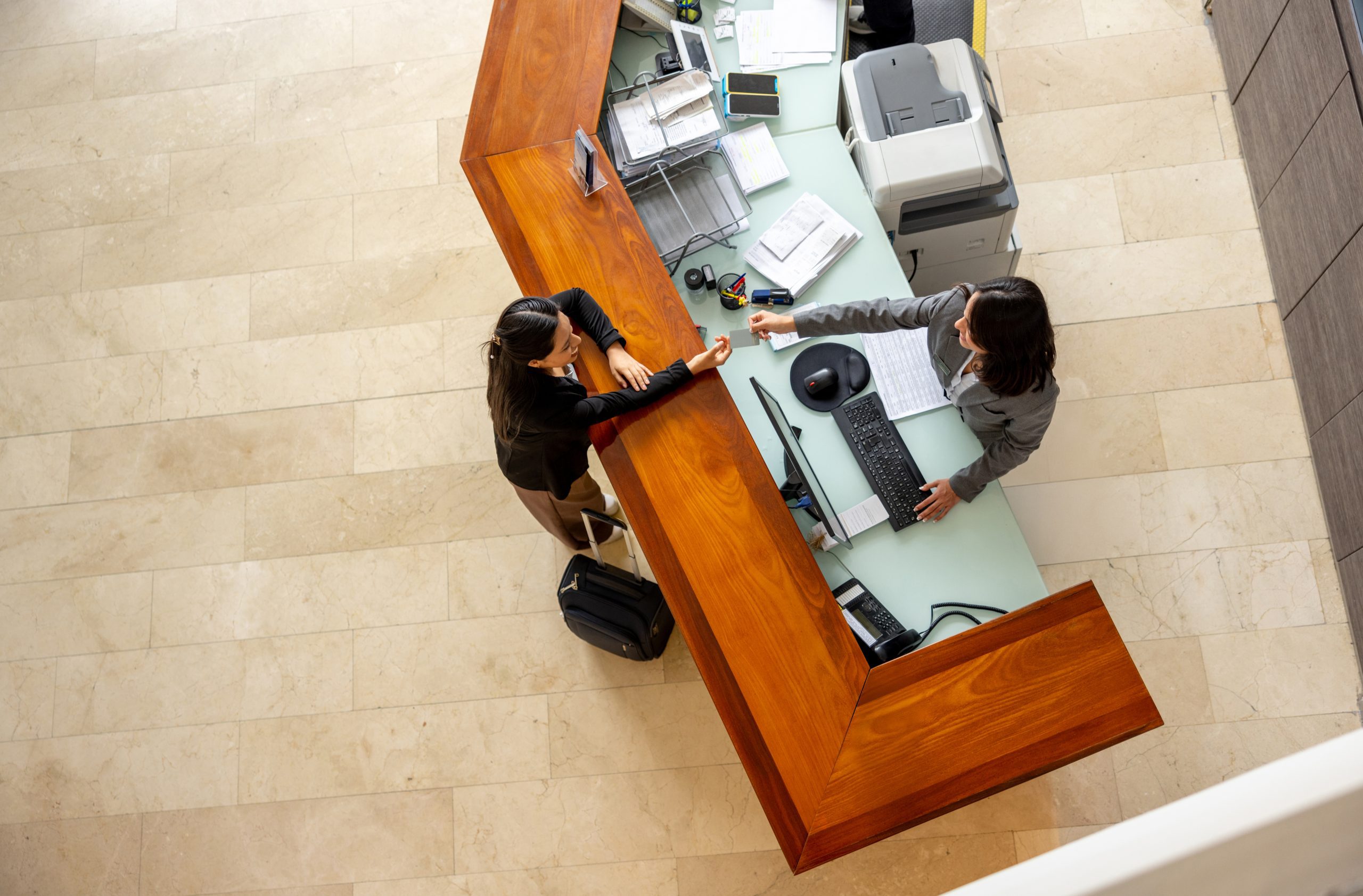 Overhead view of two women interacting at a modern reception desk with a light-colored top and wood paneling.